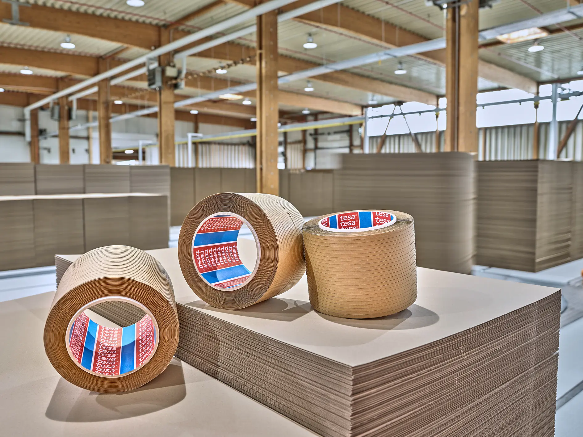 Three rolls of brown packing tesa tape with red labels are placed on stacks of large, flat sheets of cardboard. The setting appears to be a spacious industrial warehouse with wooden beams and high ceilings. (This text has been generated by AI)