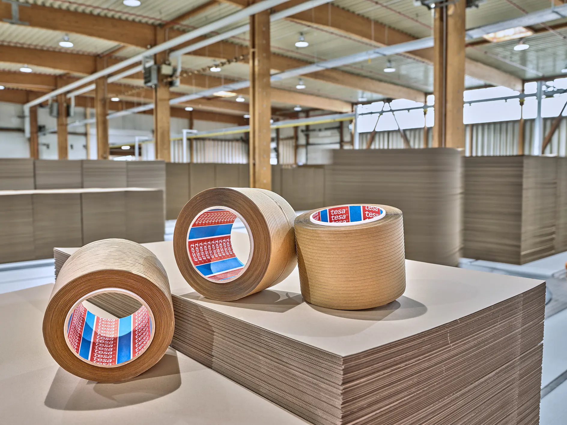 Three rolls of brown packing tesa tape with red labels are placed on stacks of large, flat sheets of cardboard. The setting appears to be a spacious industrial warehouse with wooden beams and high ceilings. (This text has been generated by AI)