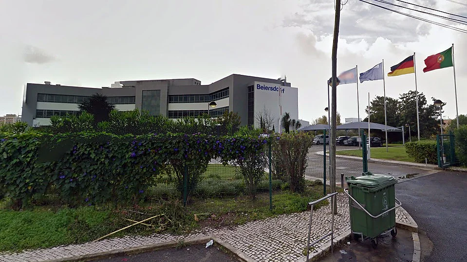 A modern building with the Beiersdorf logo is seen behind a lush green hedge. Four flags are flying on poles in front of the building, next to a path with a green trash bin by the roadside. The sky is overcast. (This text has been generated by AI)