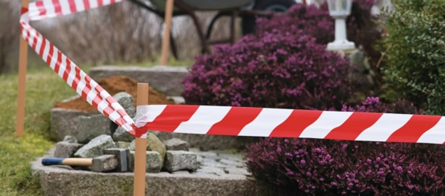 Red and white caution tesa tape cordons off an area with stone steps in a garden. There are purple shrubs nearby, and various landscaping tools such as a shovel and a trowel are visible on the ground. (This text has been generated by AI)