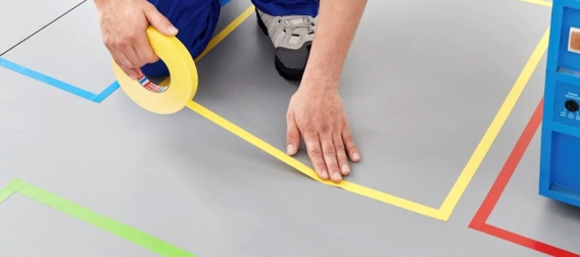 A person wearing blue pants and gray shoes is applying yellow tesa tape on a gray floor within a marked geometric area with colored lines. A blue object is partially visible on the right side of the image. (This text has been generated by AI)