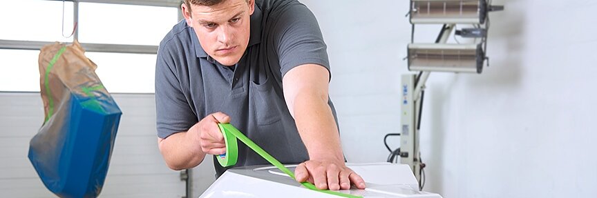 Một người đàn ông mặc áo sơ mi xám đang dán băng keo tesa màu xanh lá cây vào một vật thể lớn màu trắng trong xưởng. Một vật phẩm được che phủ treo gần đó và một lò sưởi đứng ở phía sau bên cạnh một cánh cửa kiểu gara. (This text has been generated by AI)