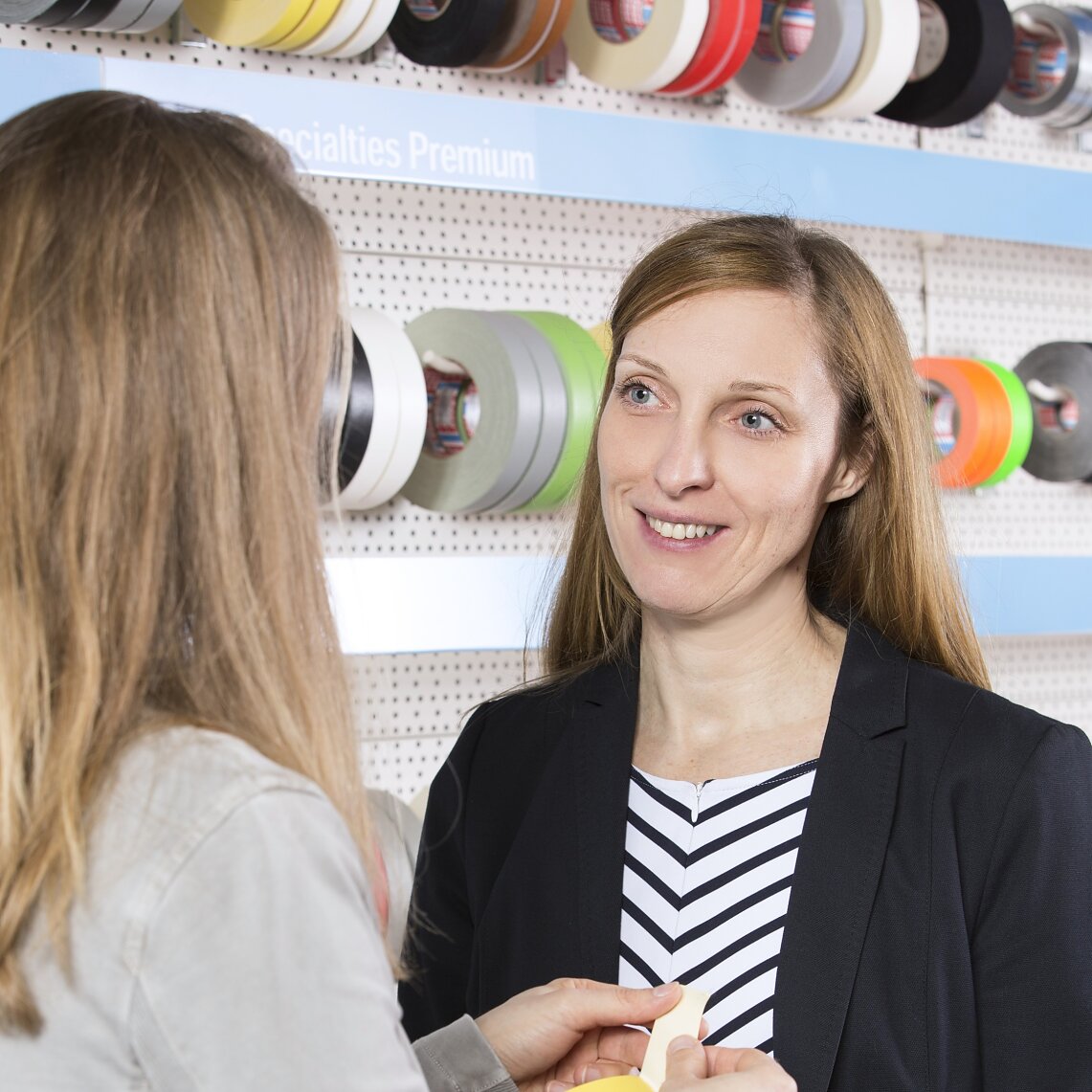 Two women are standing in front of a display of colorful tesa tape on a pegboard. One woman, in a striped shirt and blazer, is facing the camera, while the other, with long hair, is turned away, holding some samples. (This text has been generated by AI)