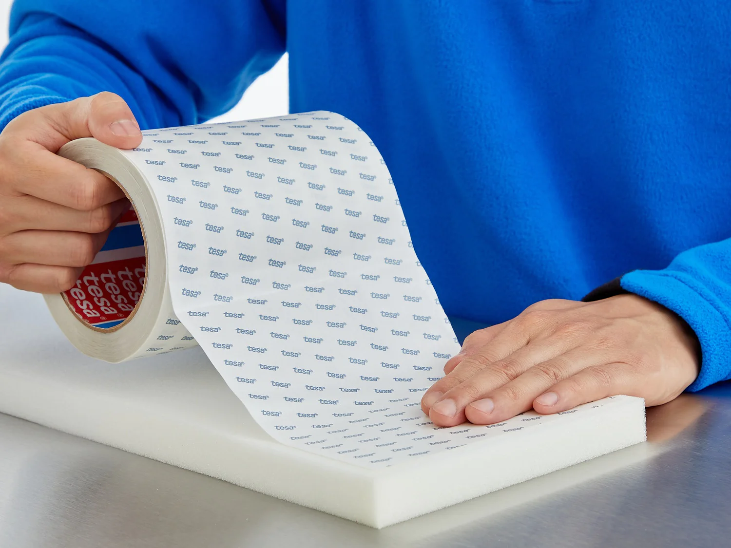 A person in a blue shirt is unrolling and applying a strip of tesa tape with a printed pattern onto a white foam surface. The persons left hand holds the tesa tape roll, while the right hand smooths it onto the foam. (This text has been generated by AI)