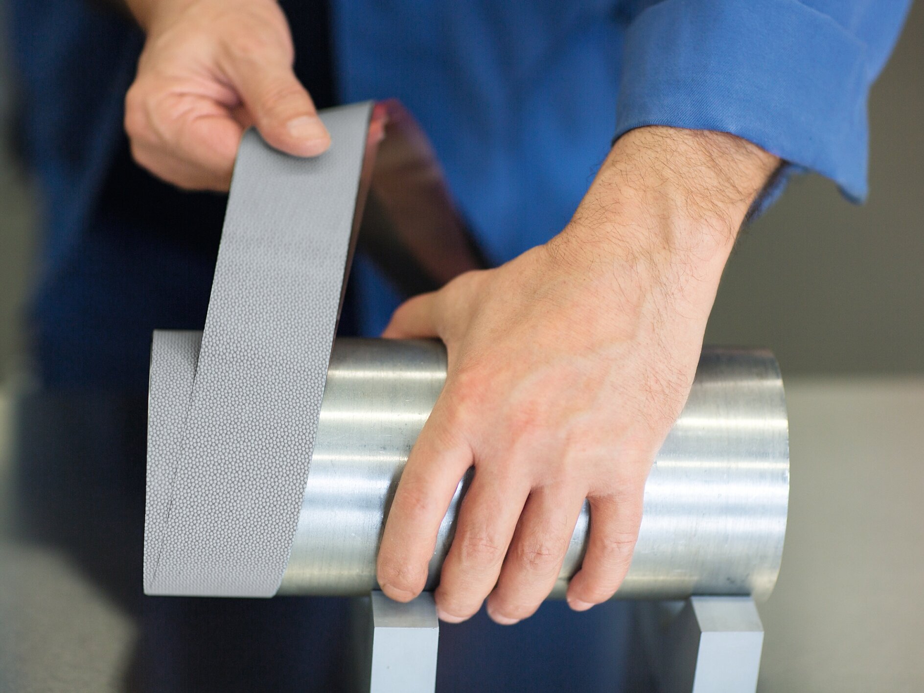 A person in a blue shirt demonstrates applying a strip of metallic, textured tesa tape to a cylindrical steel object. The object is placed on two gray stands on a smooth surface. (This text has been generated by AI)