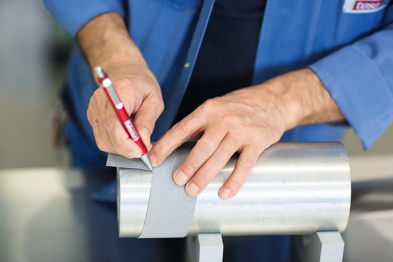 A person in a blue shirt is marking a cylindrical metal object with a red pencil while using tesa tape, which has a gray gauze-like appearance. The object rests on a stand, and the person appears focused on precise measurements or adjustments. (This text has been generated by AI)