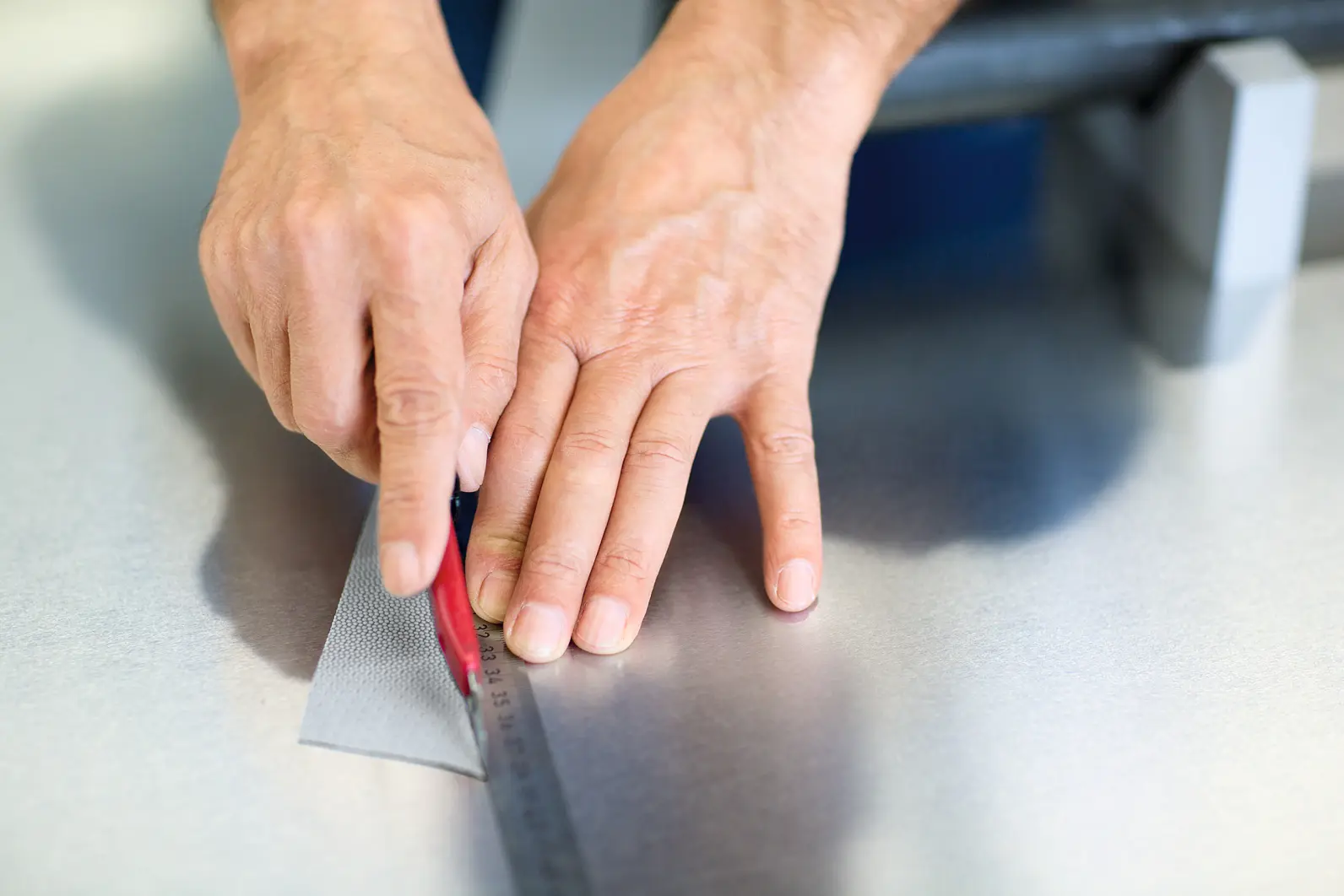 Hands cutting a piece of material with a red utility knife along a metal ruler on a smooth surface, using tesa tape. (This text has been generated by AI)