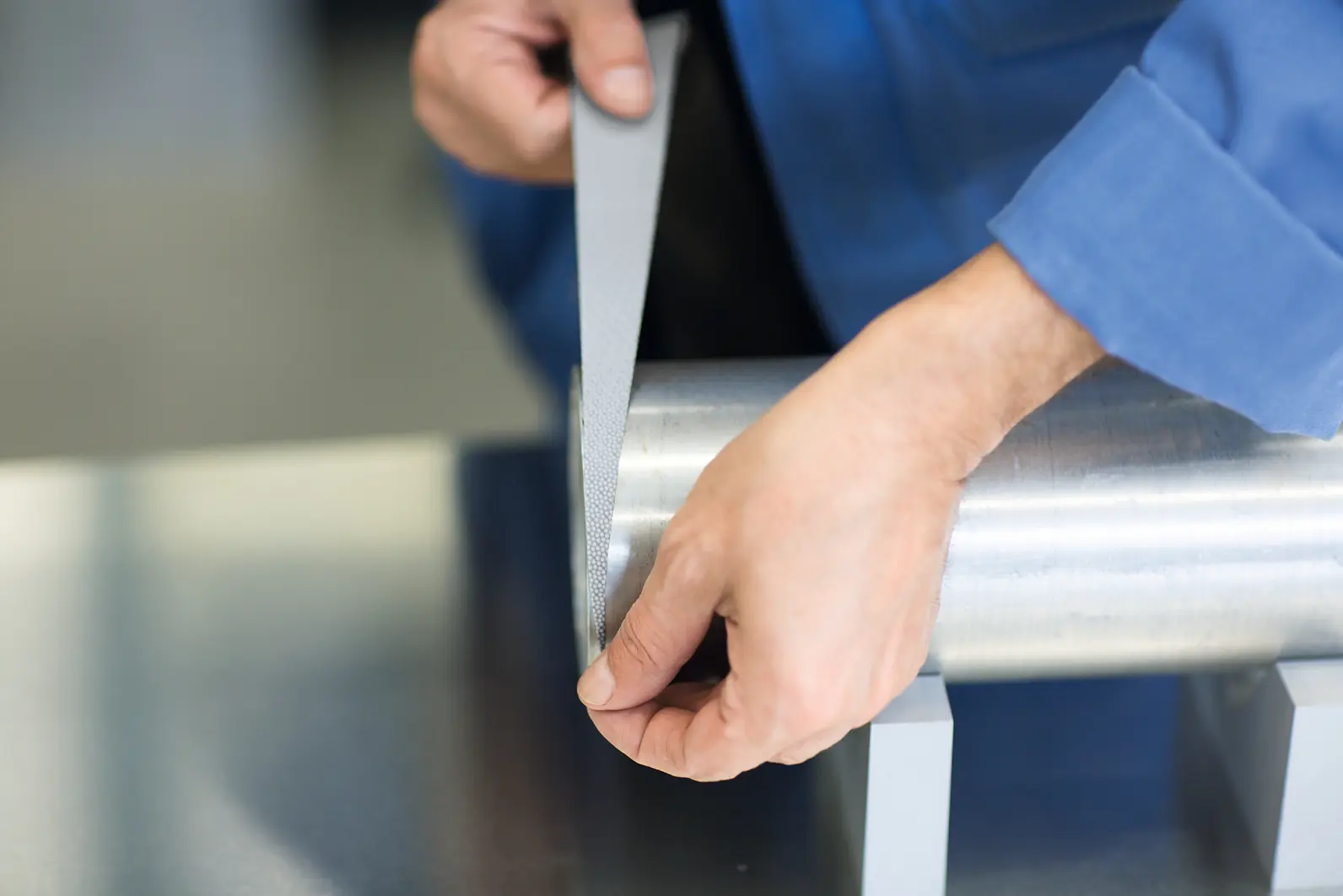 A person in a blue shirt uses a metal file to smooth the surface of a cylindrical metal object, which is supported by two small blocks. The image focuses on the persons hands and the tool in a workshop setting. (This text has been generated by AI)
