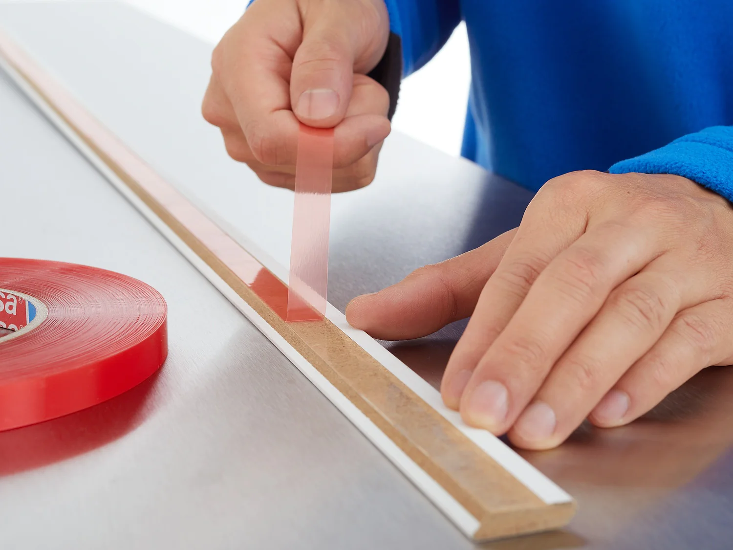 A person wearing a blue sleeve applies red double-sided adhesive tesa tape to a long, thin white strip on a metal surface. A red roll of tesa tape is positioned beside them. (This text has been generated by AI)