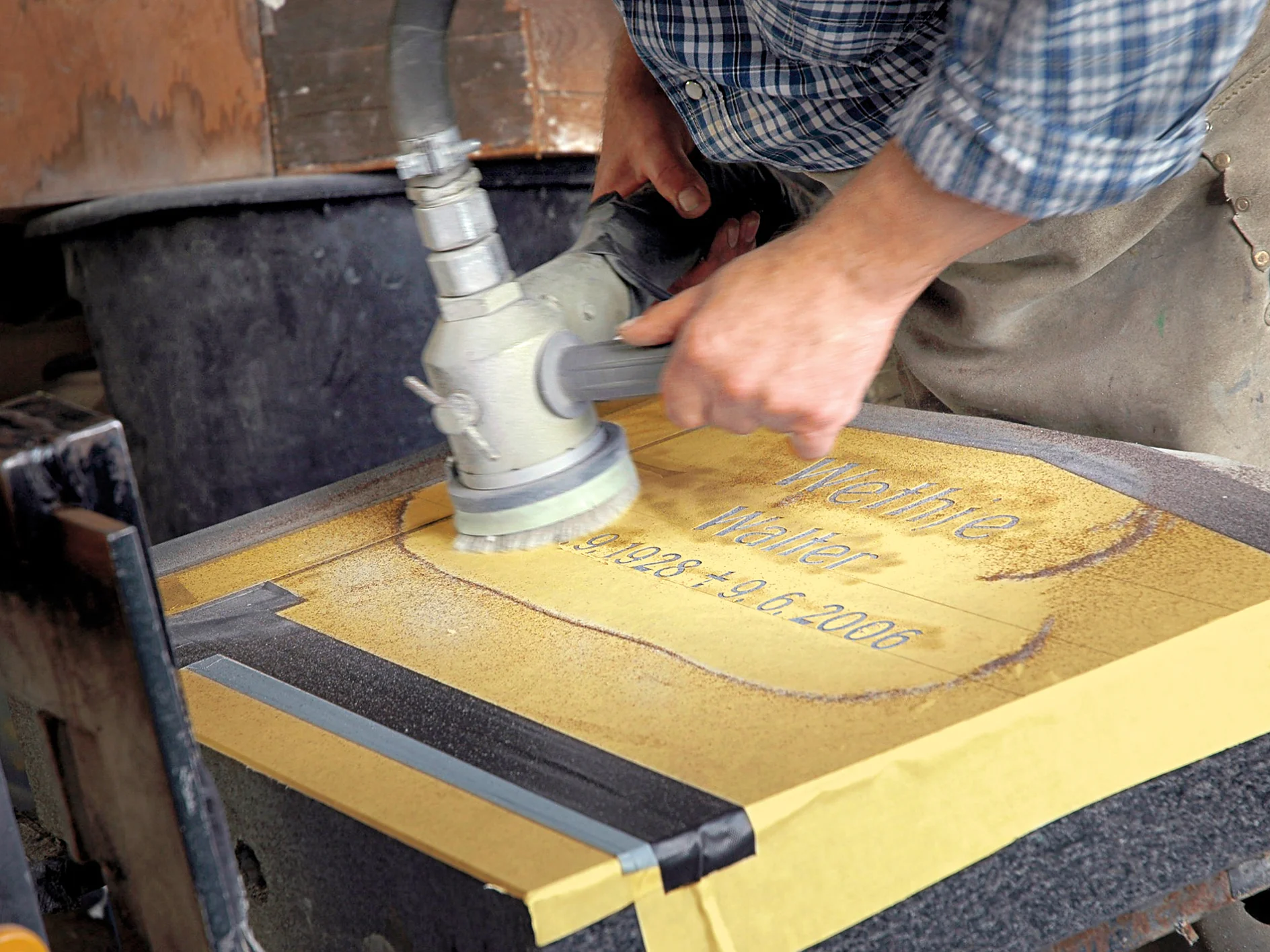 A person using a power tool to sand or polish a stone surface with the words Meinhte Walter, 9.1.1924 - 9.6.2006 visible on it. The stone is secured around the edges with tesa tape, and the person is wearing a plaid shirt and apron. (This text has been generated by AI)