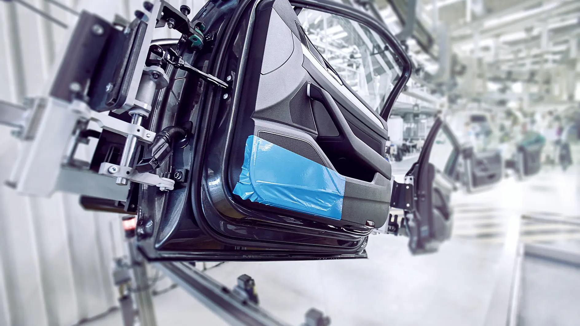 A car door is mounted on an assembly line in an industrial setting. The interior side of the door is visible, partially covered with blue protective tesa tape. The background shows more equipment and blurred parts of the assembly line. (This text has been generated by AI)