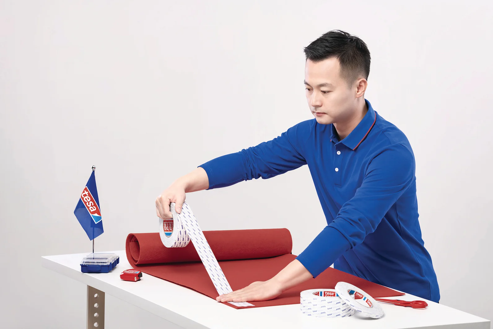 Một người mặc áo sơ mi xanh đang dán băng keo tesa lên một tấm vải đỏ đã được cuộn trên một chiếc bàn trắng. Gần đó, có một lá cờ nhỏ, một thước dây và những cuộn băng keo tesa khác. (This text has been generated by AI)