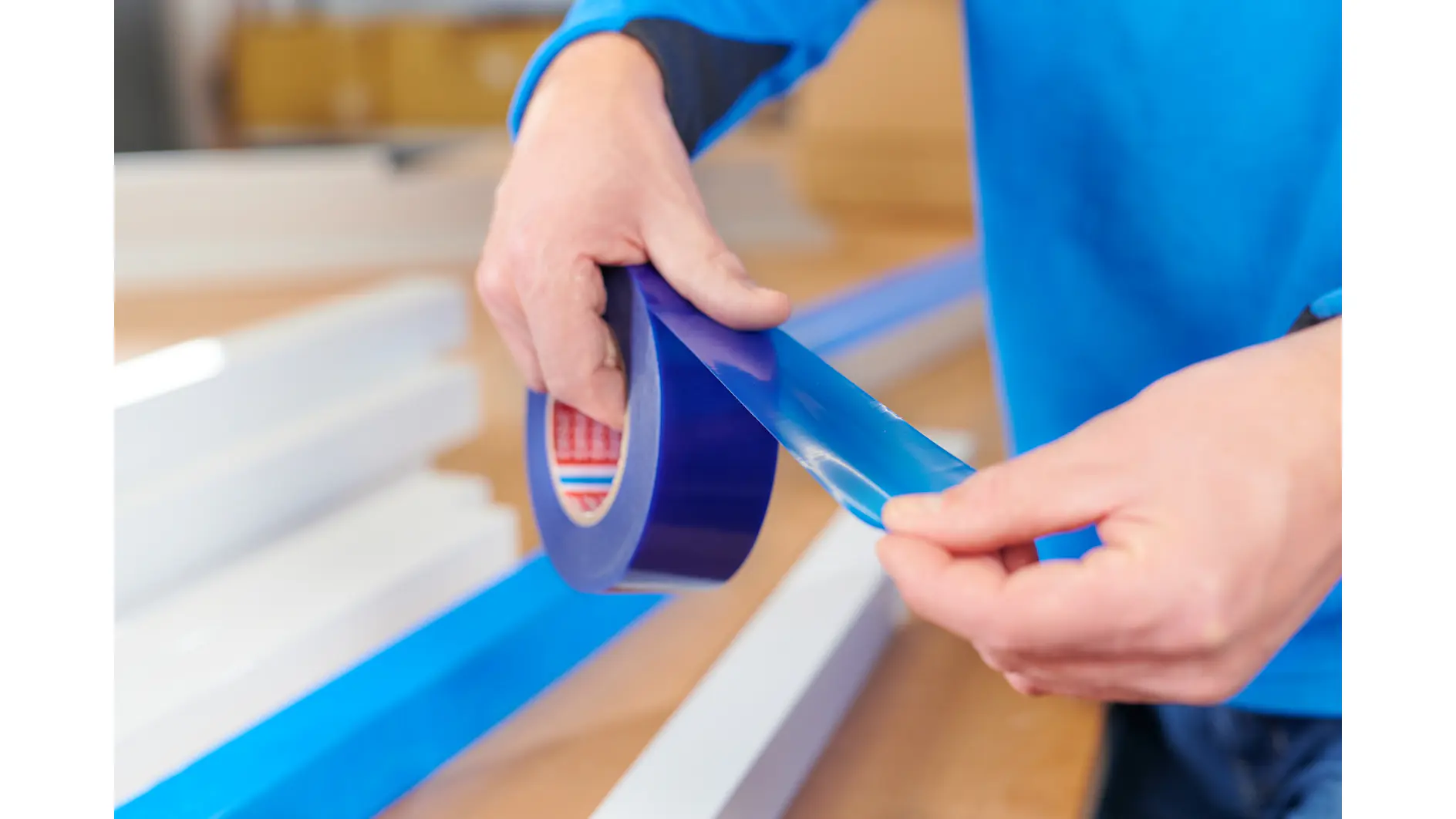 A person in a blue shirt is unrolling a piece of blue tesa tape from a roll. The background shows white rectangular objects on a wooden surface. The hands are positioned to tear the tesa tape. (This text has been generated by AI)