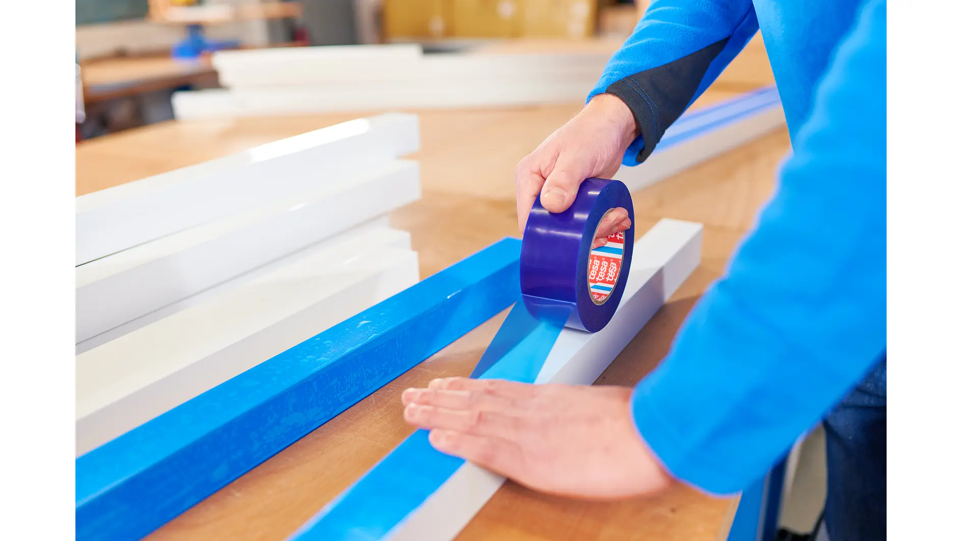 A person wearing a blue sweater applies blue tesa tape to a long, narrow piece of white material on a wooden table. Various similar white and blue materials are scattered on the table. (This text has been generated by AI)