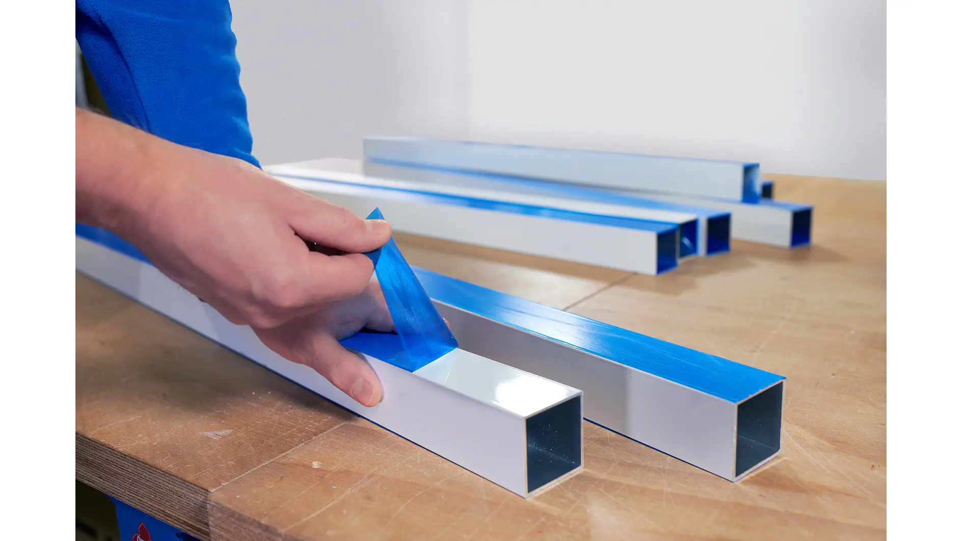 A person peels blue protective film from a white rectangular metal tube on a wooden table. Several similar tubes in the background also have tesa tape partially removed. The person is wearing a blue sleeve. (This text has been generated by AI)