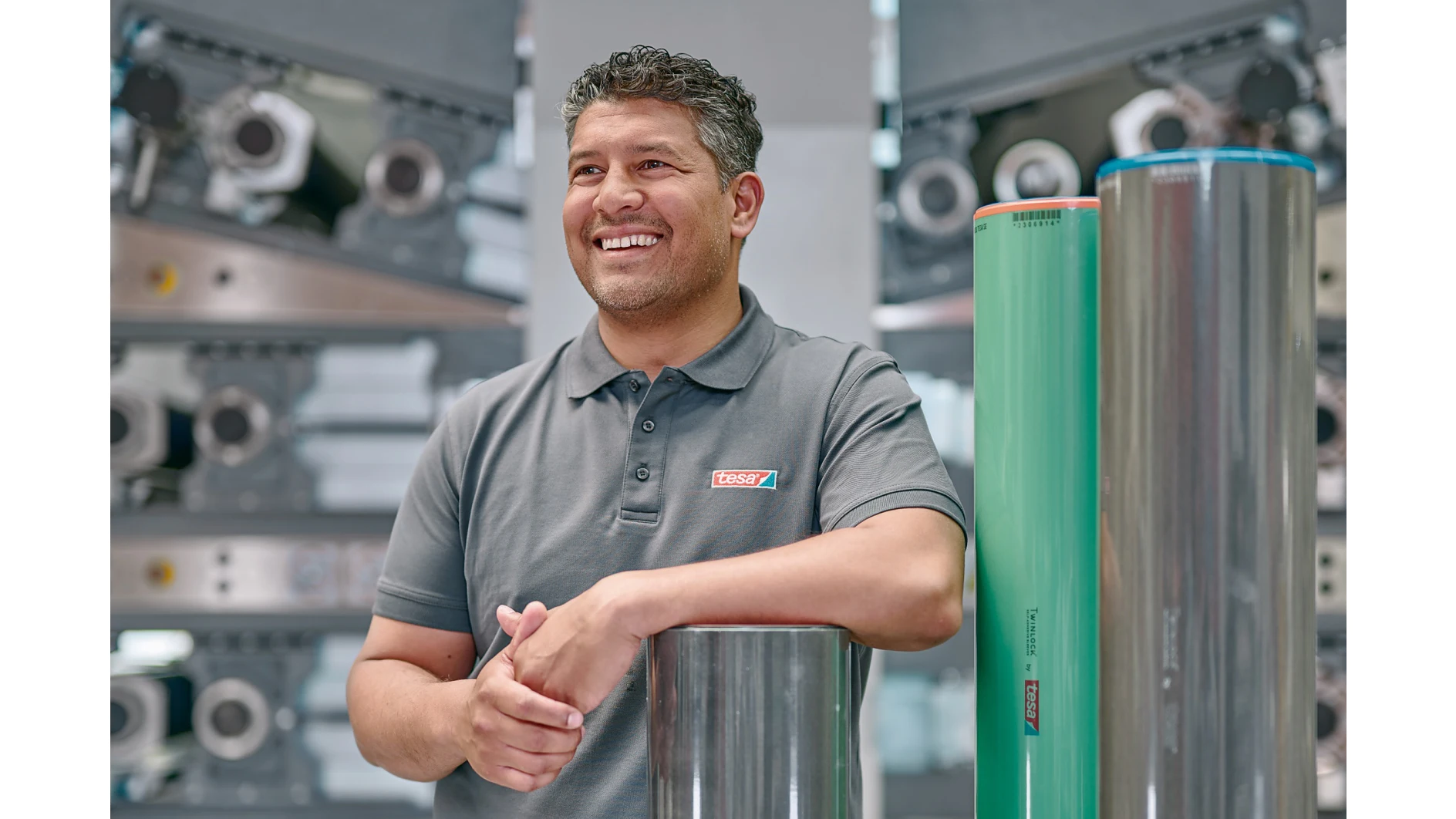 A man in a gray polo shirt stands smiling with his hands resting on a metallic cylinder in a machinery-filled environment. Two large cylindrical rolls, one metallic and one green, stand upright beside him. (This text has been generated by AI)