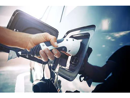 A person connects a charging cable to the charging port of an electric vehicle. The image is captured from a close-up angle, highlighting the hand gripping the cable against the cars shiny blue surface. (This text has been generated by AI)
