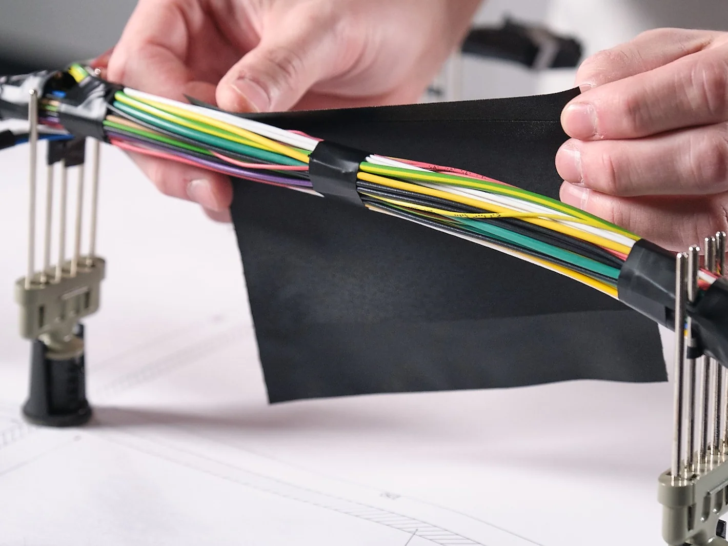 Hands are arranging a bundle of multicolored electrical wires secured with black tesa tape. The wires are positioned on a table with tools and clamps holding them in place. A dark rectangular object is held by the hands above the wires. (This text has been generated by AI)