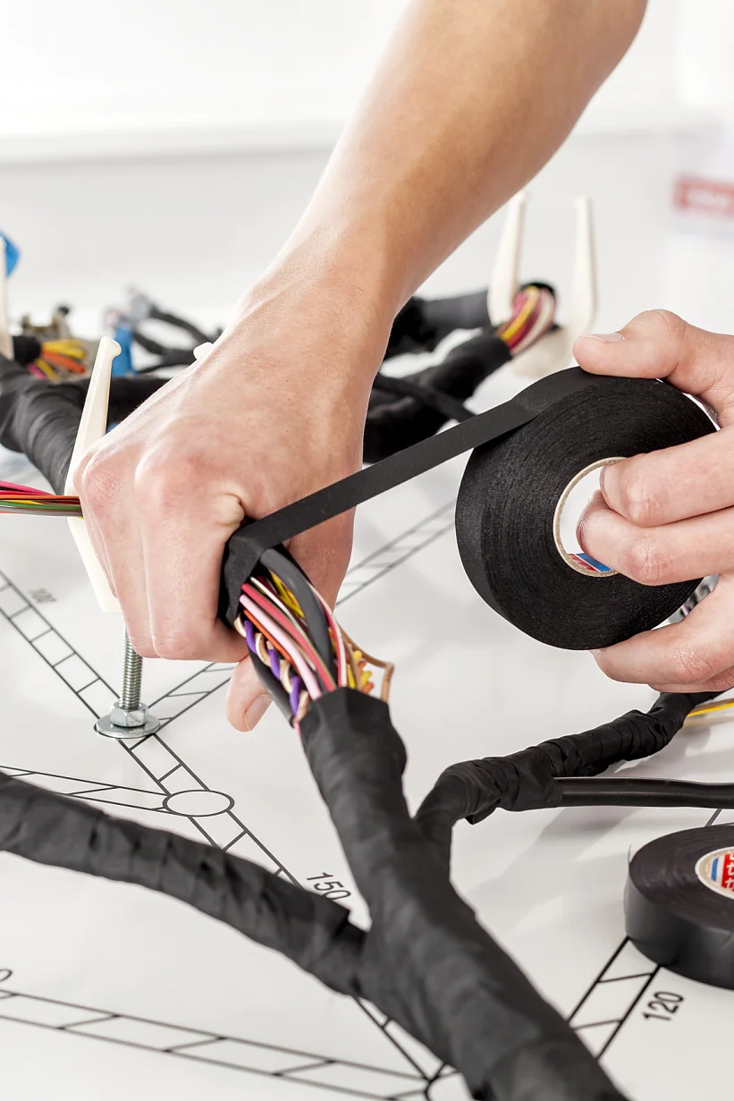 A person is wrapping tesa tape around a bundle of colored wires on a work surface. Several wire bundles are secured with clamps. Two rolls of tesa tape are visible, with one actively being used. (This text has been generated by AI)