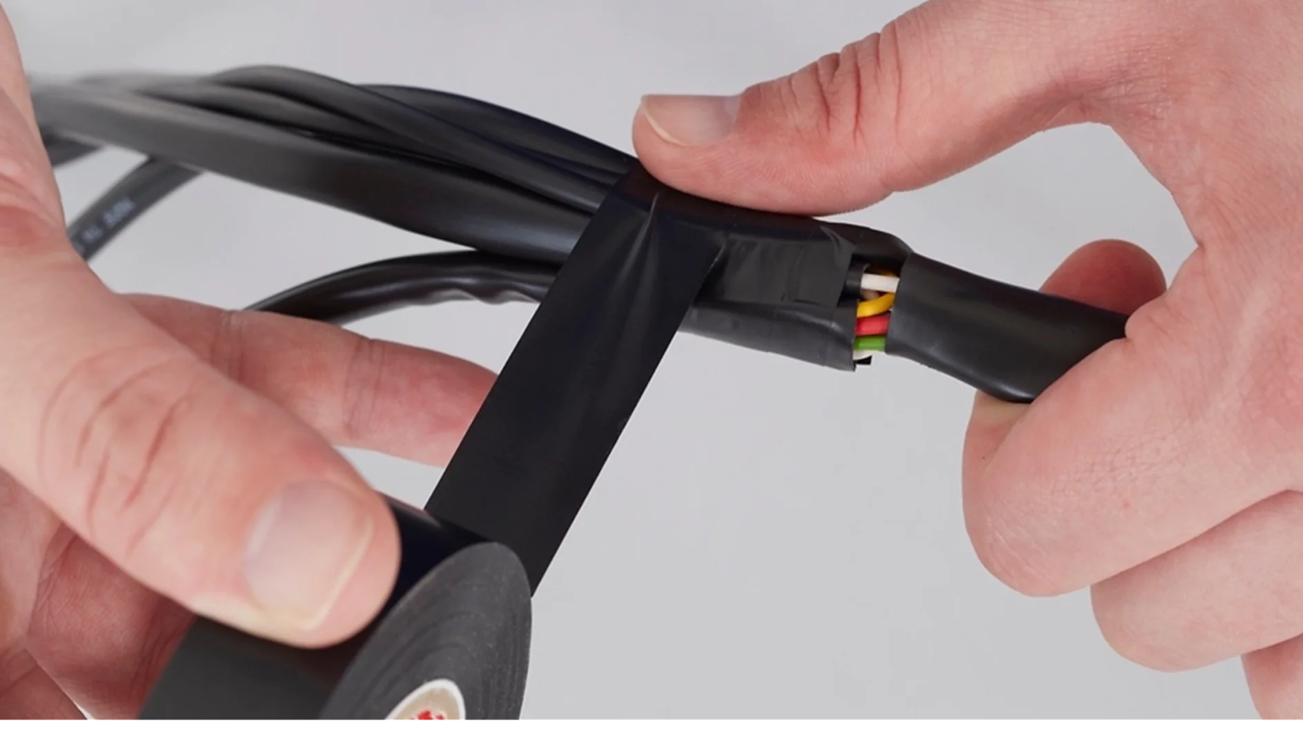 A person wraps black tesa tape around a group of colored wires. The tesa tape is being applied to secure and insulate the wires, which include green, yellow, and red colors. The background is plain white. (This text has been generated by AI)