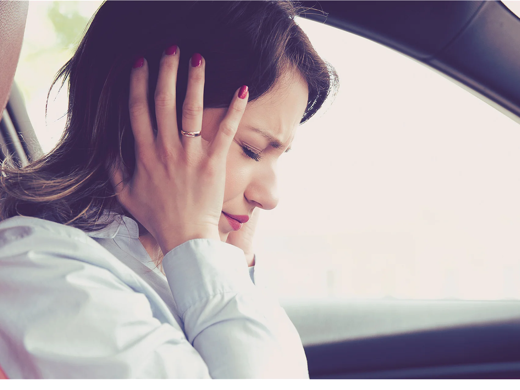 A woman is sitting in a car with both hands covering her ears. She appears to be uncomfortable or in distress. She is wearing a light-colored blouse and has dark hair. The interior of the car features dark and red accents. (This text has been generated by AI)