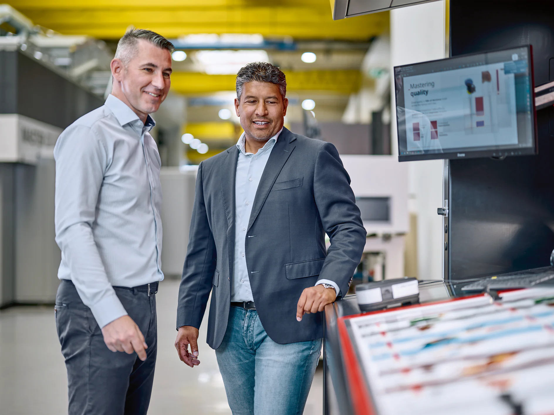 Flexo_Plate-mounting_Consultation_Bobst2023_001a-30197_V1 Two men in business attire stand in an industrial setting. One man is pointing at a computer screen displaying data, while the other smiles. They are surrounded by machinery and equipment, including several rolls of tesa tape. (This text has been generated by AI)