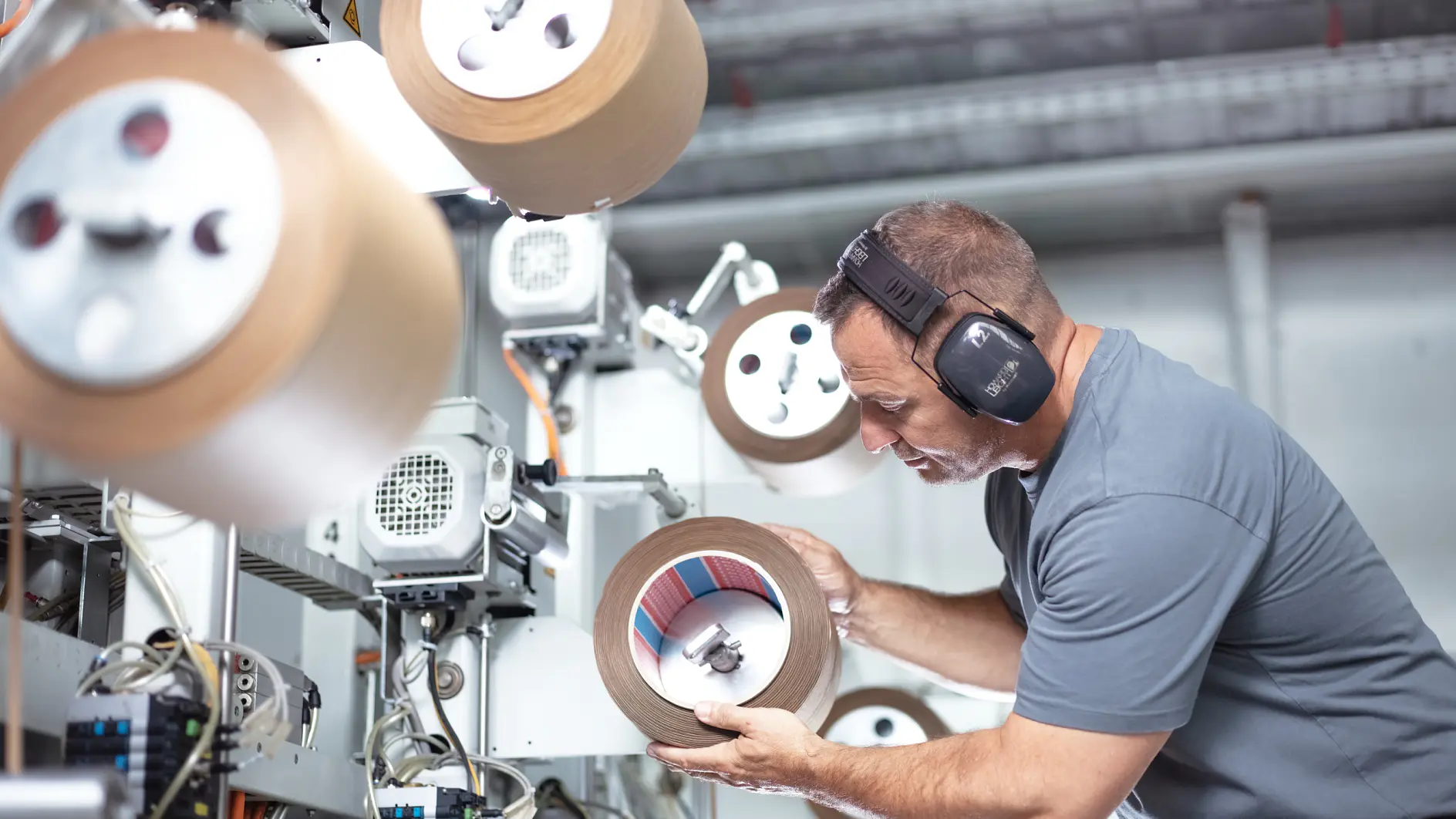 A man wearing ear protection inspects a large roll of tesa tape in an industrial setting. He is surrounded by machinery and other rolls of tesa tape, focusing intently on the one in his hands. The environment appears to be a manufacturing facility. (This text has been generated by AI)
