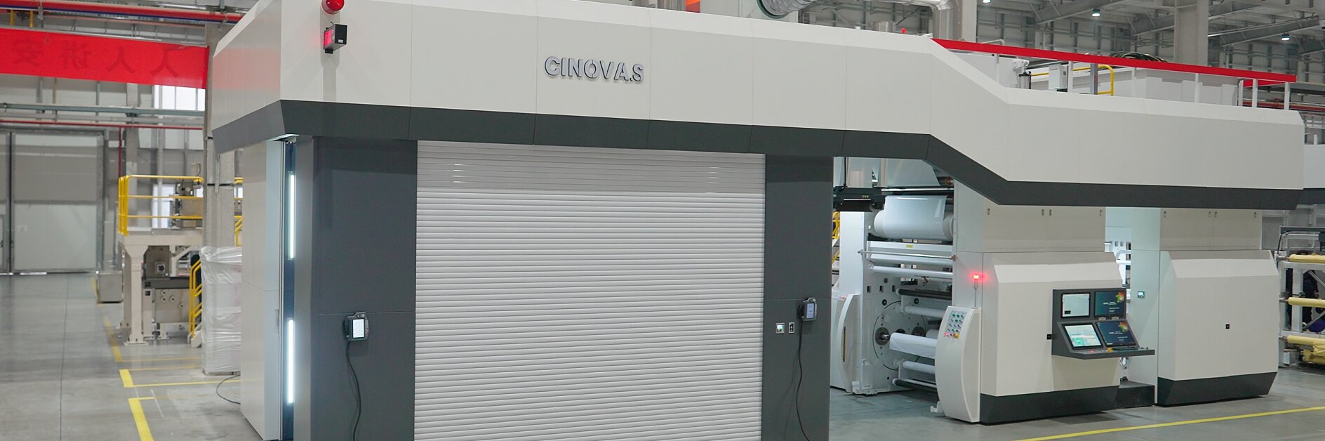 Large industrial printing machine in a factory setting. The machine is gray and white with control panels and display screens. The floor is concrete, and the area is well-lit with overhead lights. Yellow lines are marked on the floor for guidance using tesa tape.