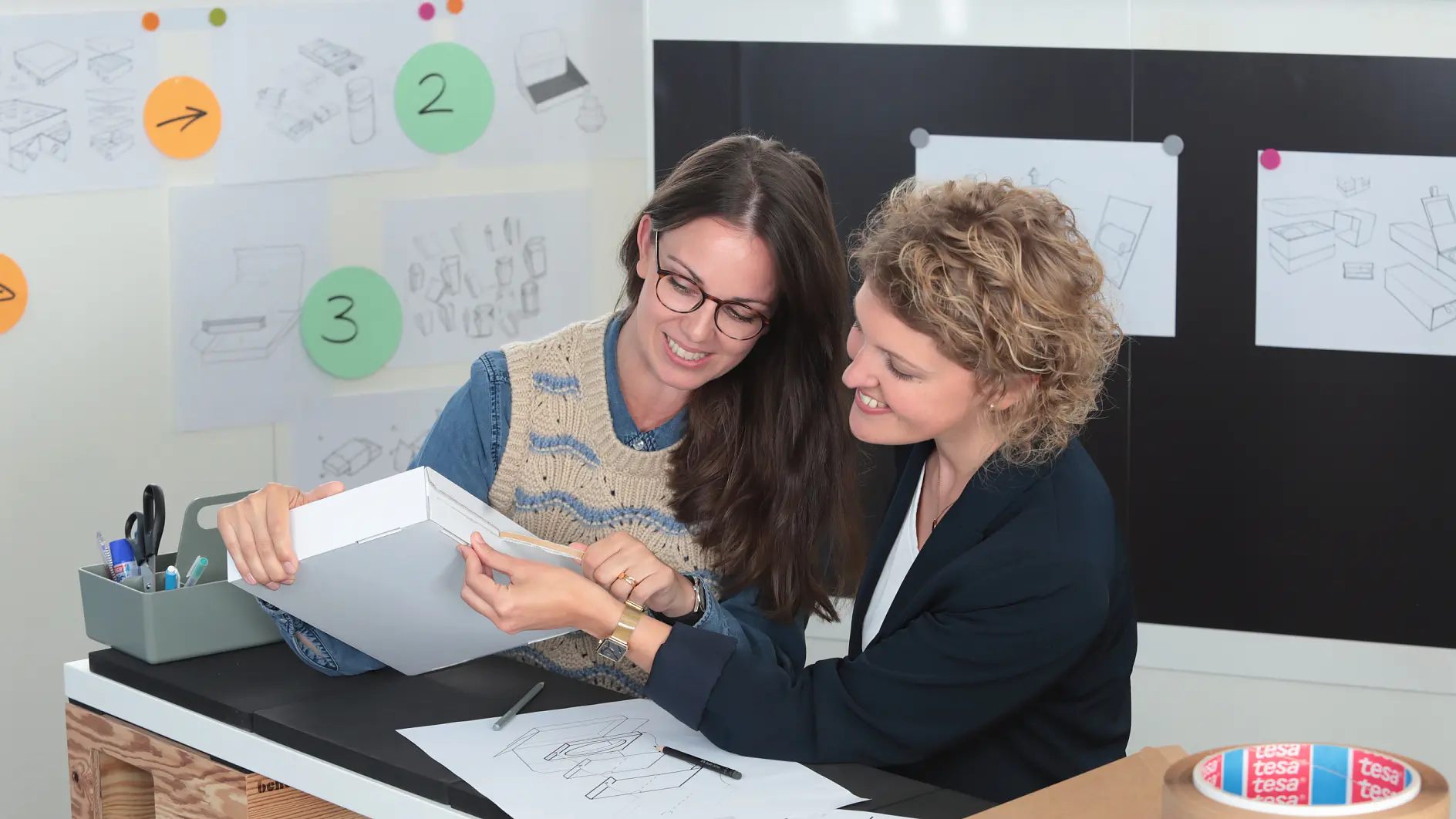 Two people are seated at a table with design sketches, examining a white box. The workspace has sketches and notes on the wall. Various office supplies, including tesa tape and markers, are on the table. Both appear engaged in discussion.