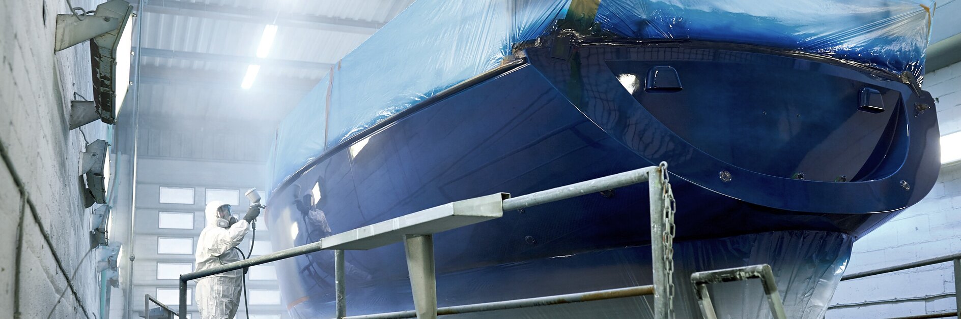 workers in protective suits spray coating on a blue boat hull inside an industrial painting booth with bright overhead lights tesa (This text has been generated by AI)