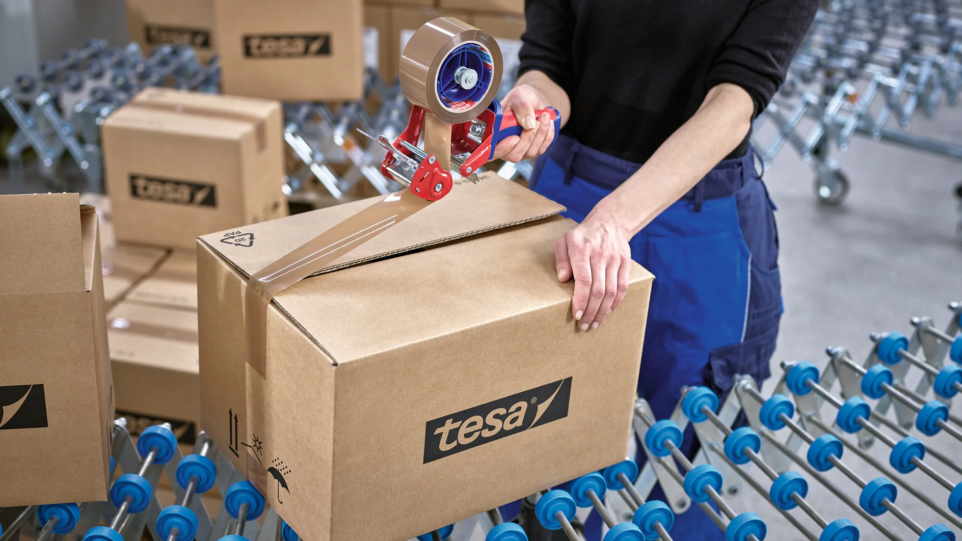 A person seals a cardboard box with a roll of packing tesa tape. The box has the tesa logo on it. Several similar boxes are on a conveyor belt in the background. The person is wearing a black top and blue pants. (This text has been generated by AI)