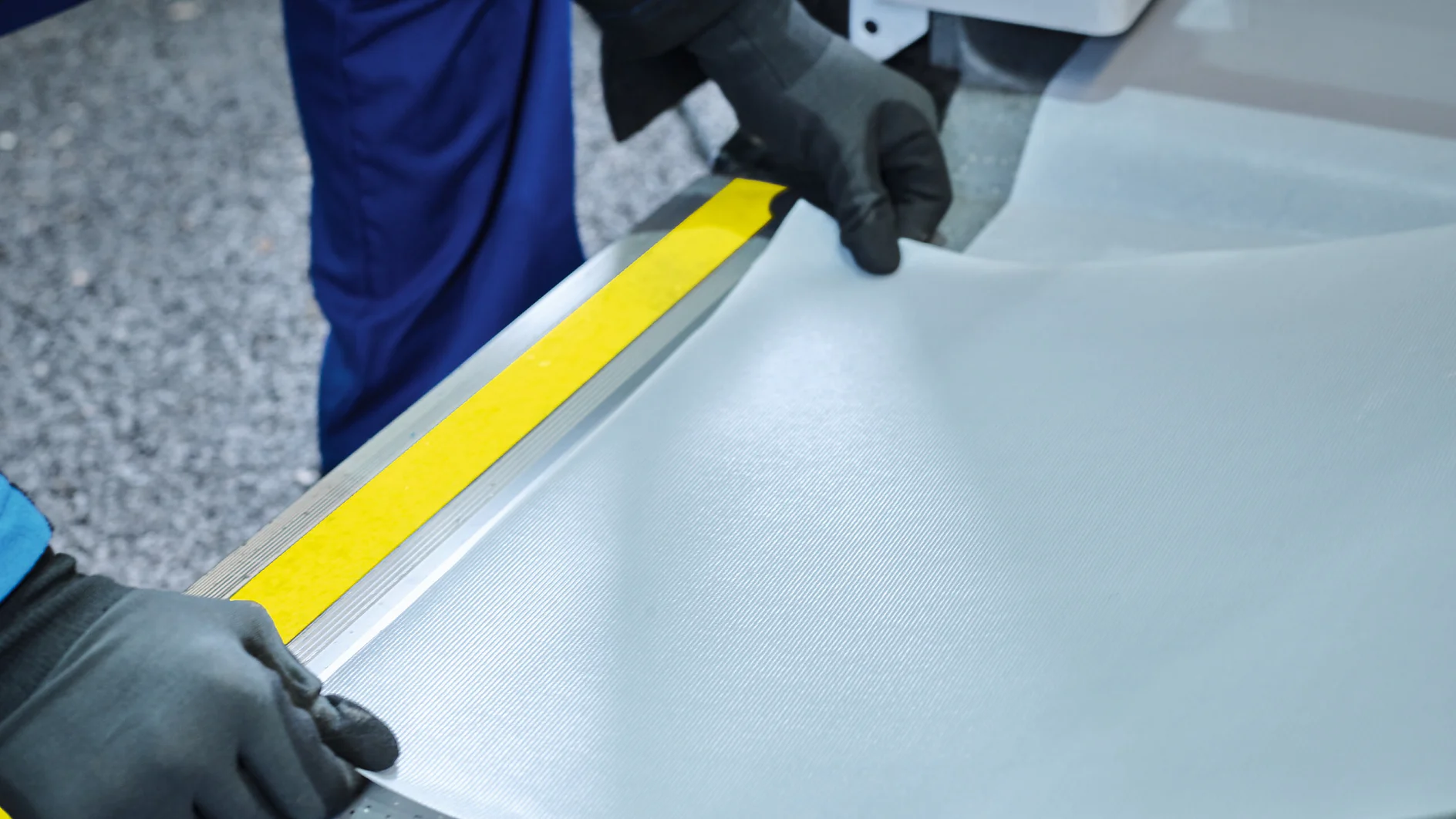 tesa-4416-anti-slip-tape-image-002-ap-cms A person wearing black gloves and a blue uniform is holding and adjusting a sheet of light gray material, possibly fabric or vinyl, next to a yellow and gray metal edge in a workspace with a textured floor. (This text has been generated by AI)
