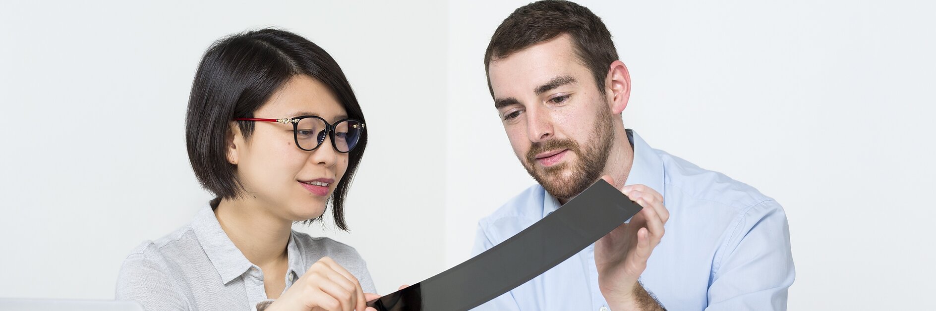 A man and woman are seated at a table in a bright room, examining a flexible, thin, black object. A laptop is open on the table, along with scattered papers. Both appear focused on the tesa tape they are holding. (This text has been generated by AI)