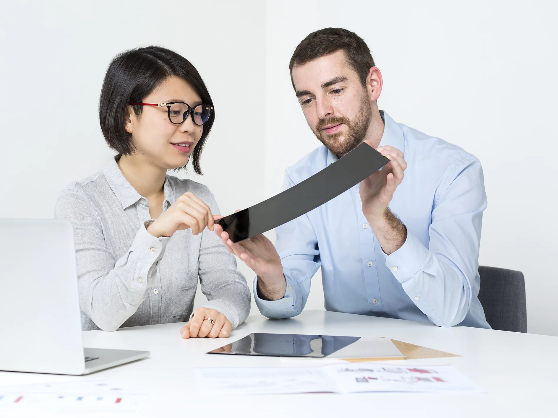 A man and woman are seated at a table in a bright room, examining a flexible, thin, black object. A laptop is open on the table, along with scattered papers. Both appear focused on the tesa tape they are holding. (This text has been generated by AI)