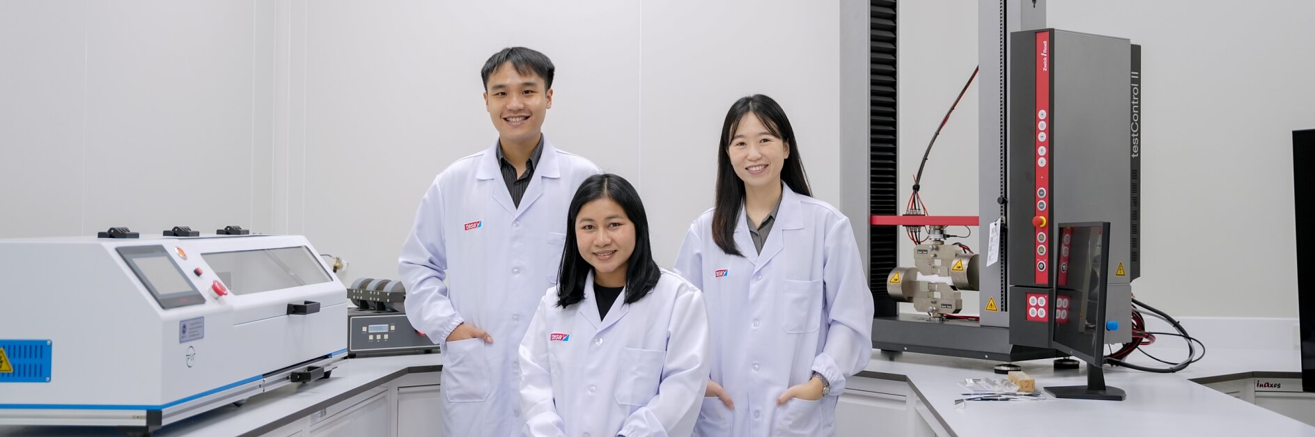 Ba người mặc áo blouse trắng đứng trong phòng thí nghiệm. Họ được bao quanh bởi các thiết bị khoa học và máy móc, bao gồm nhiều cuộn và ứng dụng của băng dính tesa, với hai người đứng và một người ngồi trên ghế, tất cả đều hướng về máy ảnh và nở nụ cười. (This text has been generated by AI)