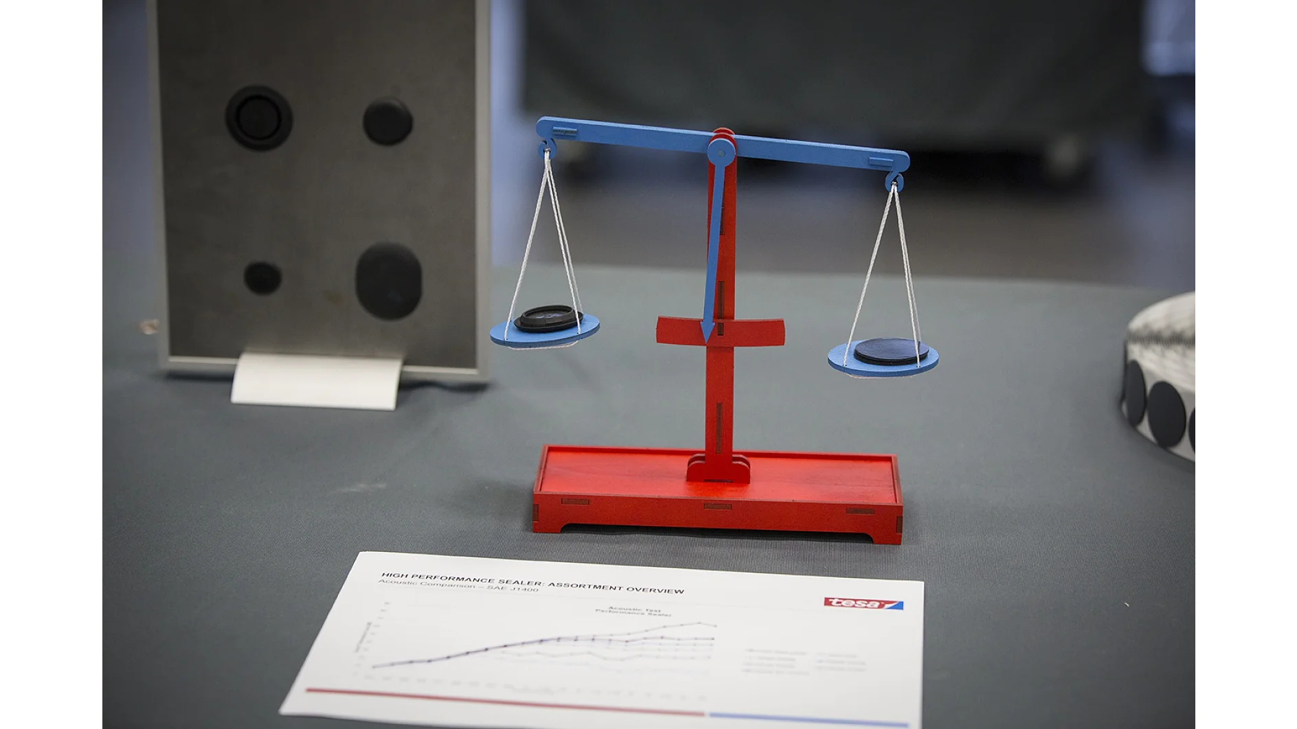 Một chiếc cân thăng bằng màu đỏ và xanh giữ một vật màu đen ở mỗi bên, cho thấy chúng bằng nhau về trọng lượng. Ở phía trước, một tờ giấy có biểu đồ và logo nằm trên bề mặt màu xám, với băng dính tesa. Các vật thể ở phía sau bị làm mờ. (This text has been generated by AI)