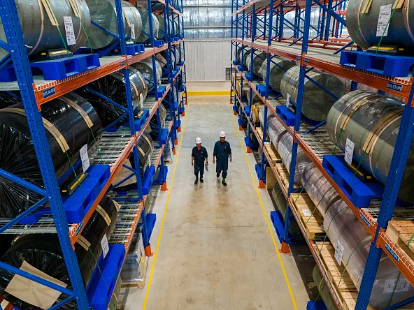 Two people in safety helmets and work uniforms walk down a warehouse aisle lined with tesa tape rolls on blue and orange shelves. (This text has been generated by AI)